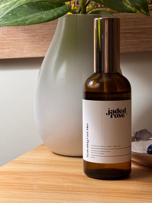 Jaded Rose Hydrating Face Mist in an amber glass bottle with a rose gold pump cap, styled on a warm timber shelf beside a grey ceramic vase with green leafy plant. 100ml.