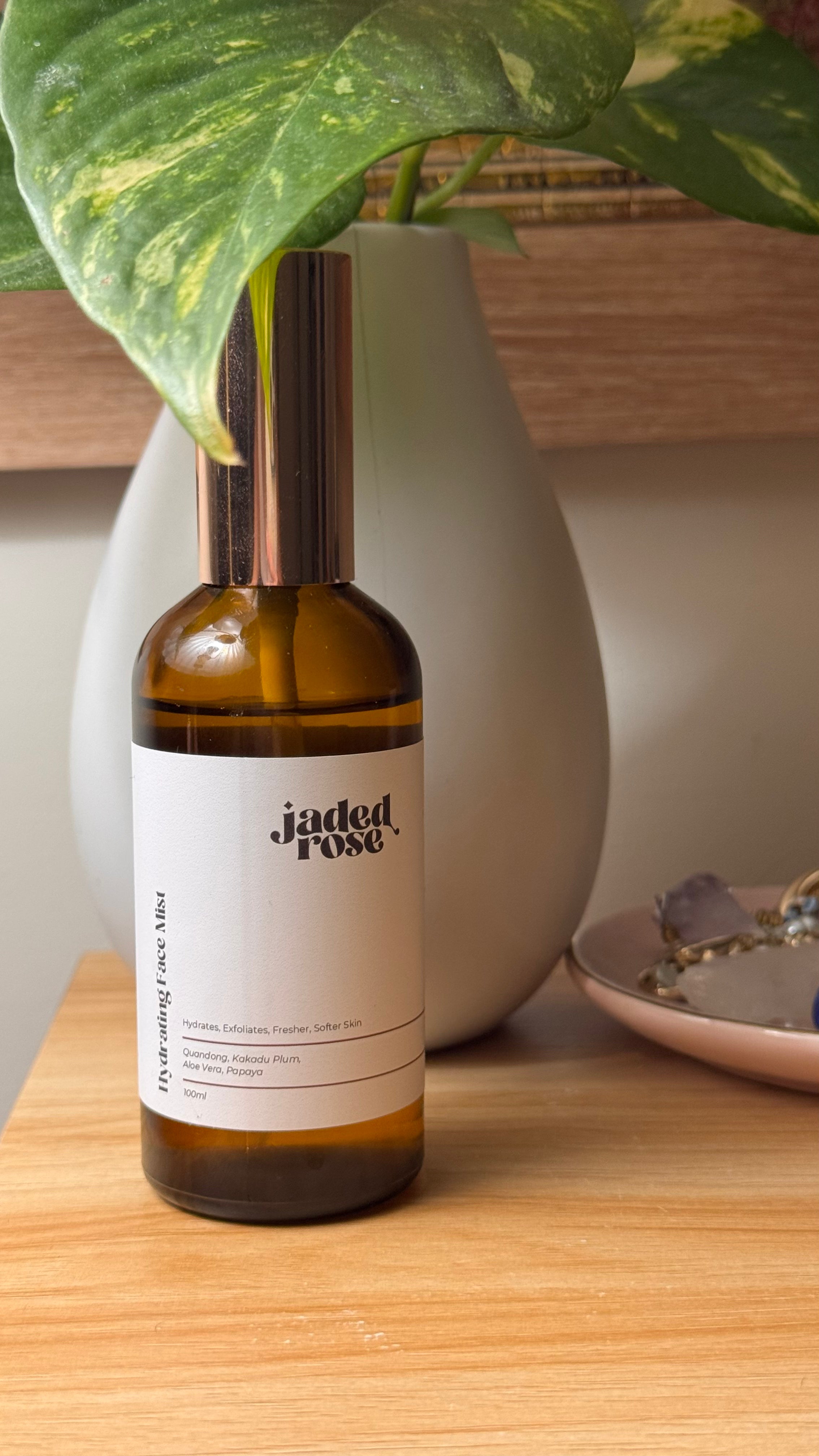 Jaded Rose Hydrating Face Mist in an amber glass bottle with a rose gold pump cap, styled on a warm timber shelf with a grey ceramic vase and trailing green and yellow pothos plant. 100ml.