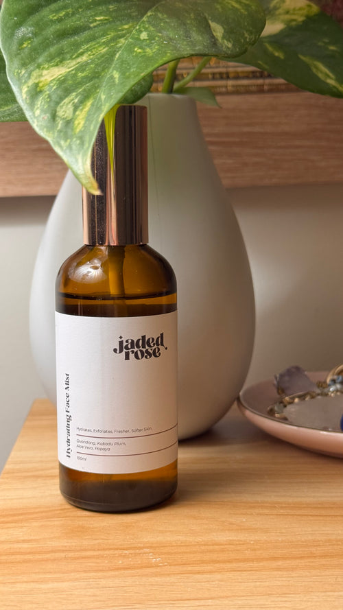 Jaded Rose Hydrating Face Mist in an amber glass bottle with a rose gold pump cap, styled on a warm timber shelf with a grey ceramic vase and trailing green and yellow pothos plant. 100ml.