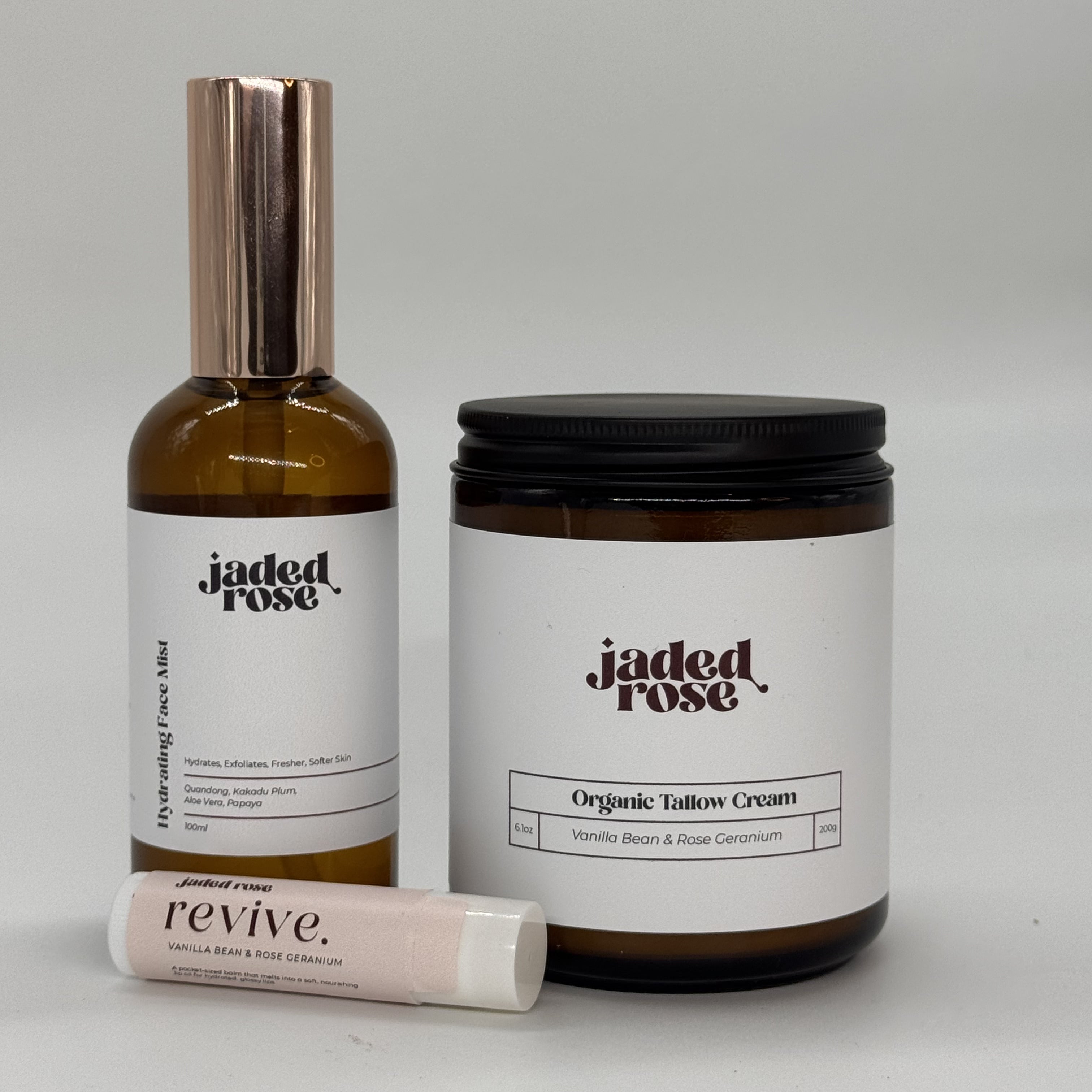 The Ritual Set by Jaded Rose, three products arranged on a light neutral surface: the Hydrating Face Mist in an amber glass bottle with a rose gold pump cap, the Organic Tallow Cream in an amber jar with a matte black lid, and the Revive Lip Balm in a blush-toned tube. All products feature the Jaded Rose logo in dark serif type on clean white labels. Vanilla Bean & Rose Geranium scent.