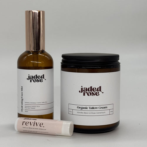 The Ritual Set by Jaded Rose, three products arranged on a light neutral surface: the Hydrating Face Mist in an amber glass bottle with a rose gold pump cap, the Organic Tallow Cream in an amber jar with a matte black lid, and the Revive Lip Balm in a blush-toned tube. All products feature the Jaded Rose logo in dark serif type on clean white labels. Vanilla Bean & Rose Geranium scent.